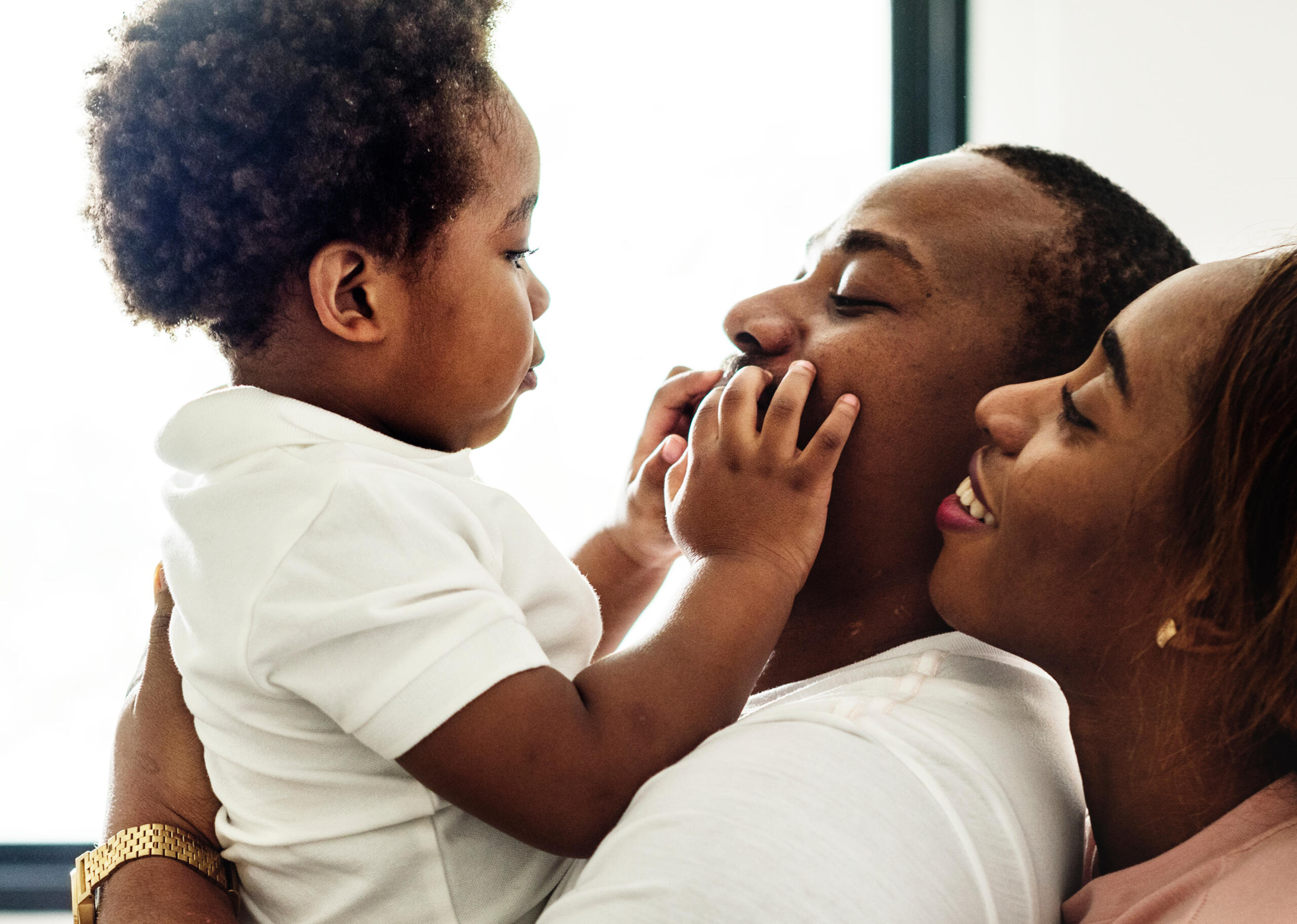 Father and mother holding the baby up while the baby is playing with the father's mouth.